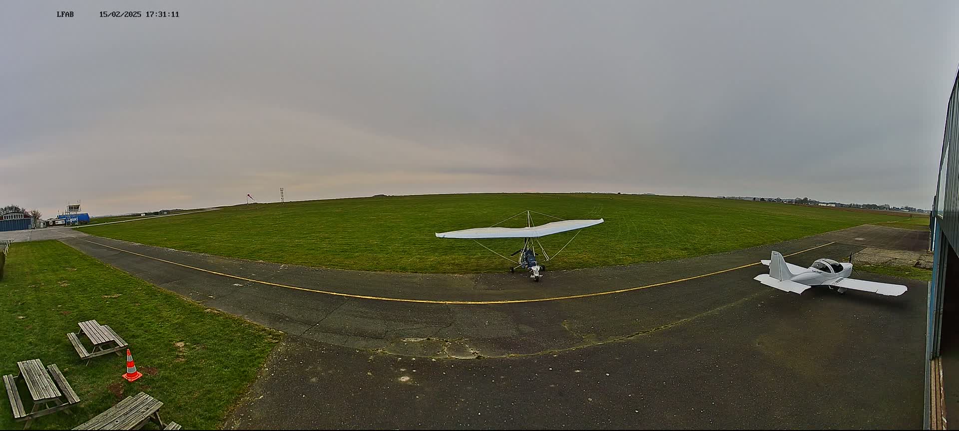A grassy airfield under an overcast sky features a hang glider and a small airplane parked on the tarmac near picnic tables and a traffic cone.