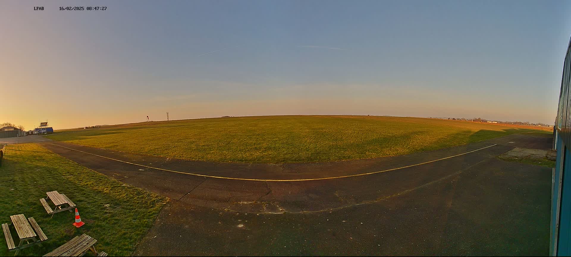A wide grassy airfield with a paved taxiway curves towards a hangar under a clear, twilight sky.