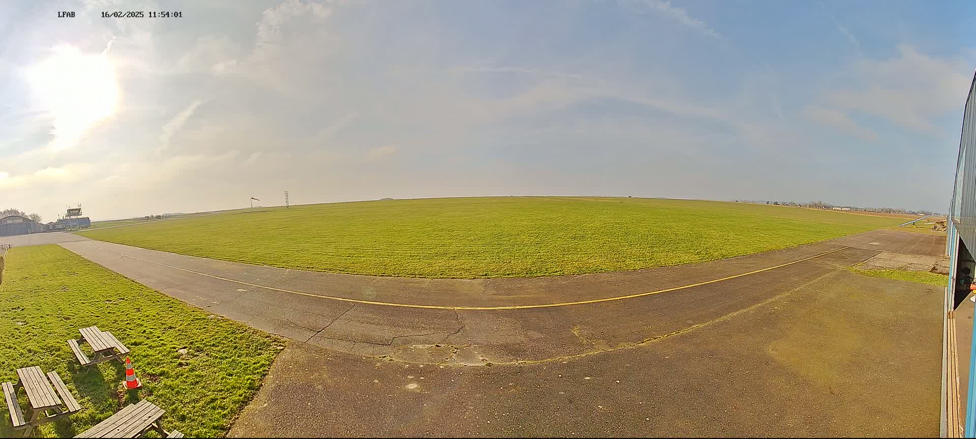 A grassy airfield with a paved runway and picnic tables is visible under a mostly sunny sky.