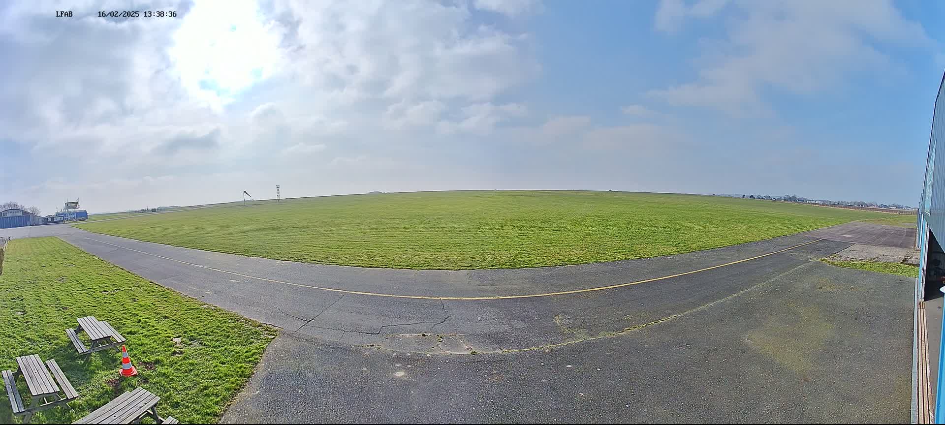 A grassy airfield with a paved taxiway curves toward a hangar under a mostly sunny sky.