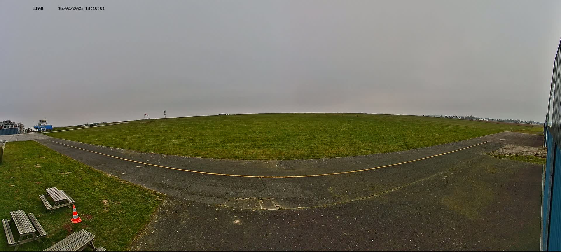 A grassy airfield with a paved taxiway and picnic tables is visible under an overcast sky.