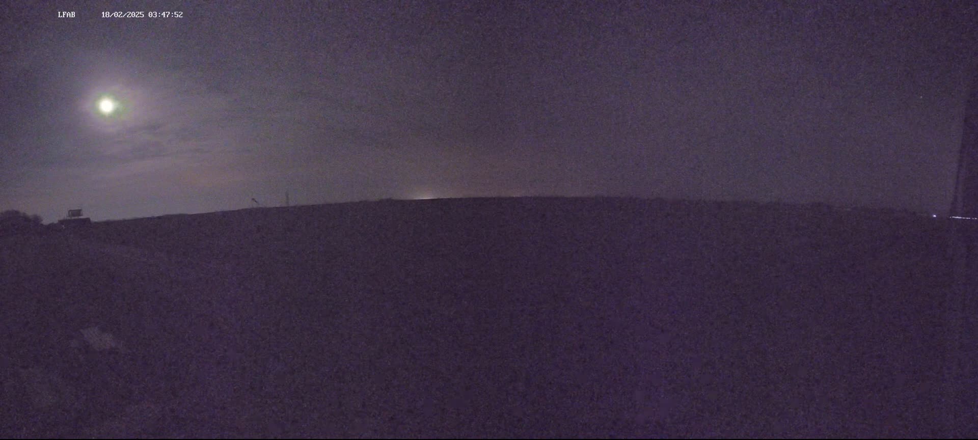 A bright moon illuminates a dark, flat landscape under a mostly clear night sky.