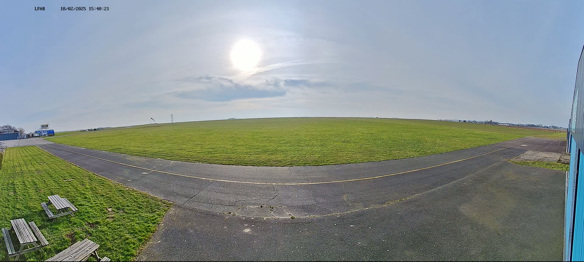 A grassy airfield with a paved taxiway, picnic tables, and a bright sun in a mostly clear sky.