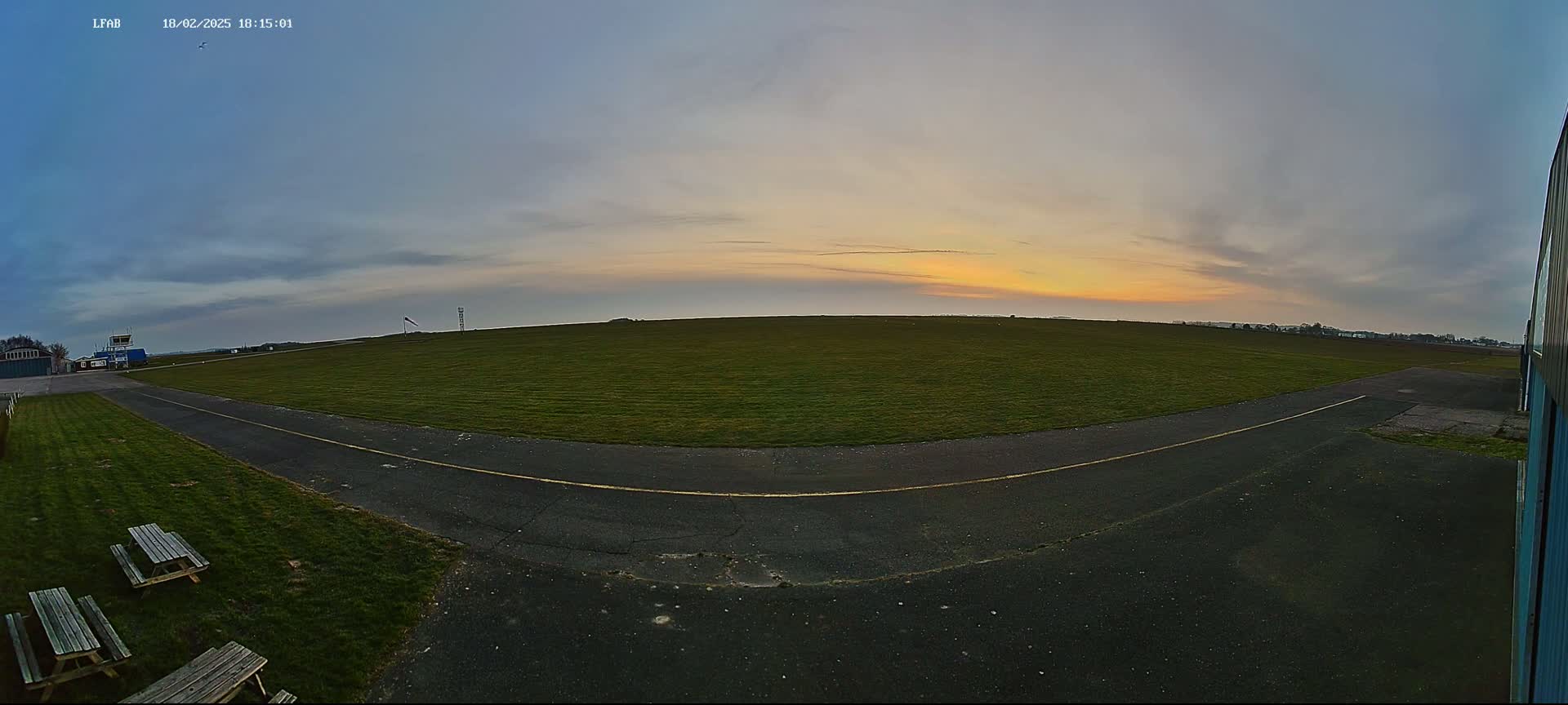 A grassy airfield at sunset, with a paved taxiway leading to a hangar, and picnic tables in the foreground.