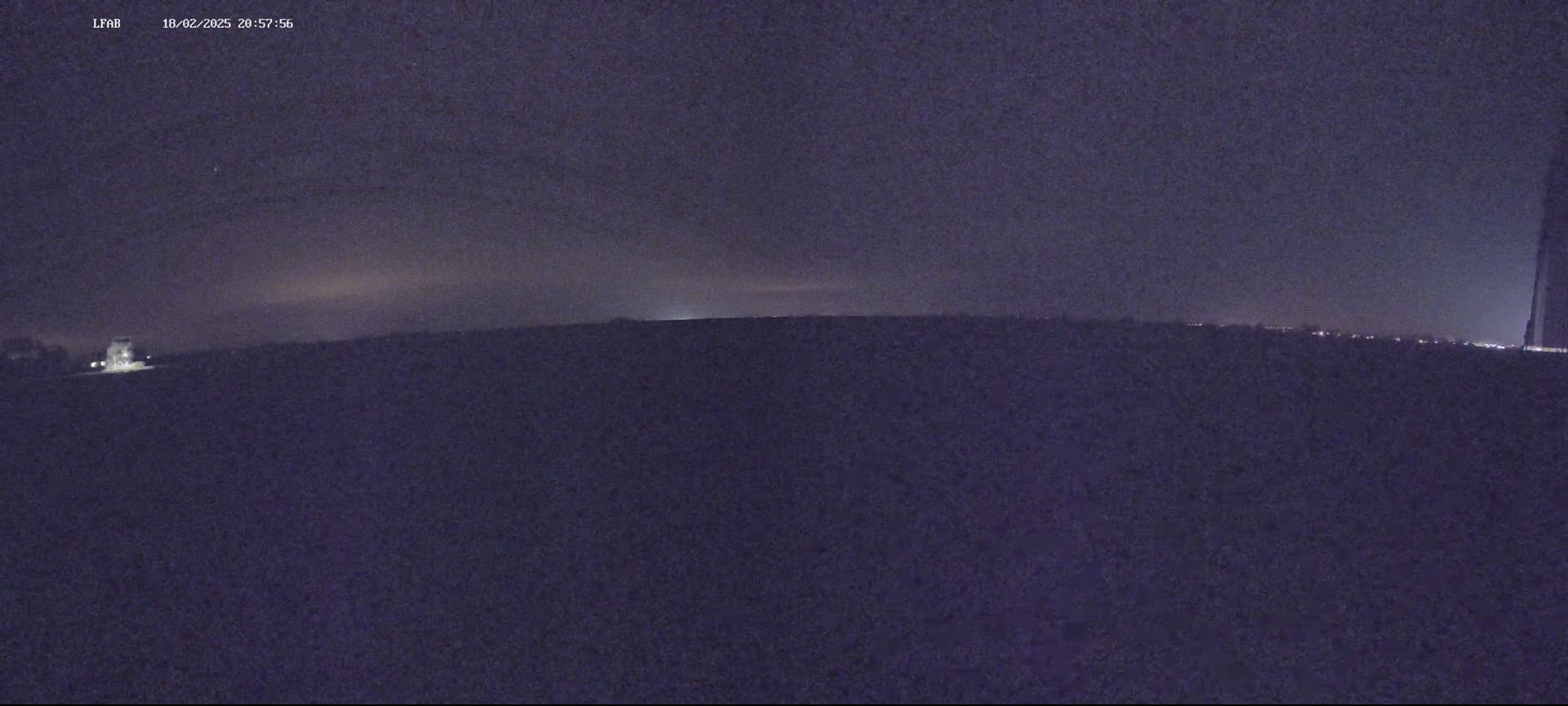A dark, nighttime landscape is shown with a dimly lit horizon and a few lights visible in the distance under a dark, cloudy sky.