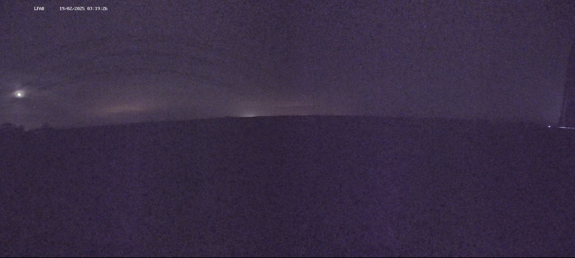 A dark, night-time scene shows a flat, dark landscape under a mostly cloudy, dusky sky with a bright light source visible in the distance.