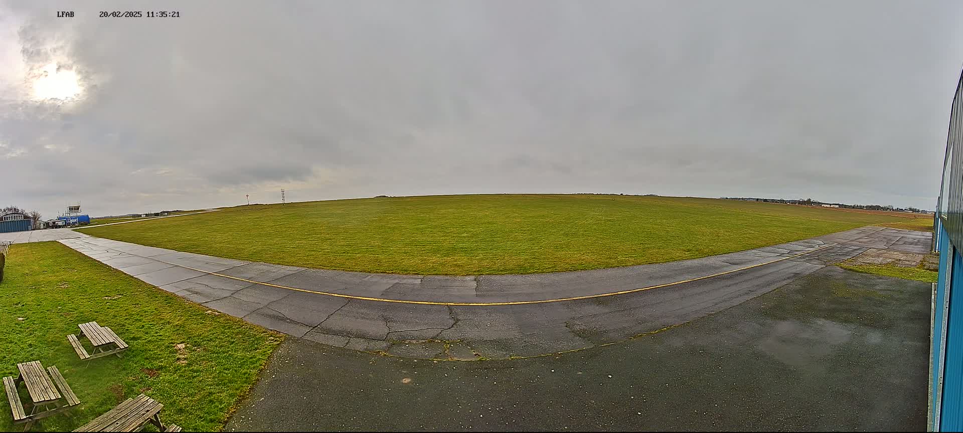 A grassy airfield under an overcast sky shows a paved taxiway curving toward a control tower and hangar.