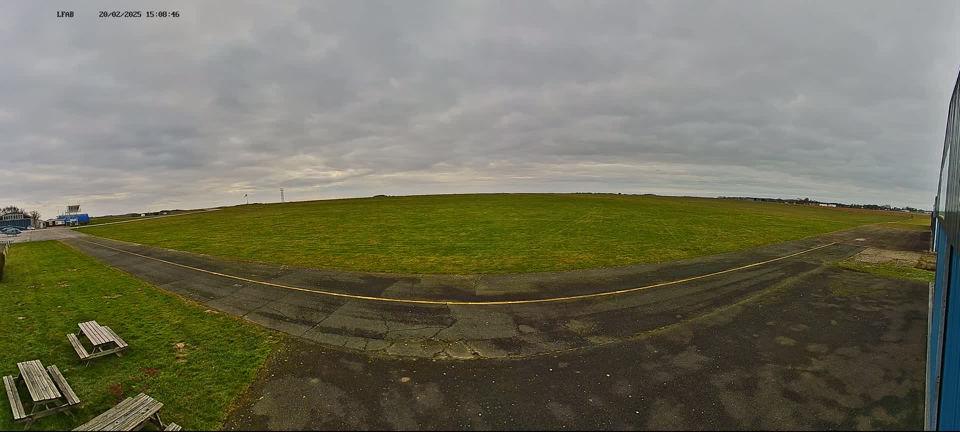 A grassy airfield under an overcast sky, with a paved taxiway curving toward a small building and picnic tables.