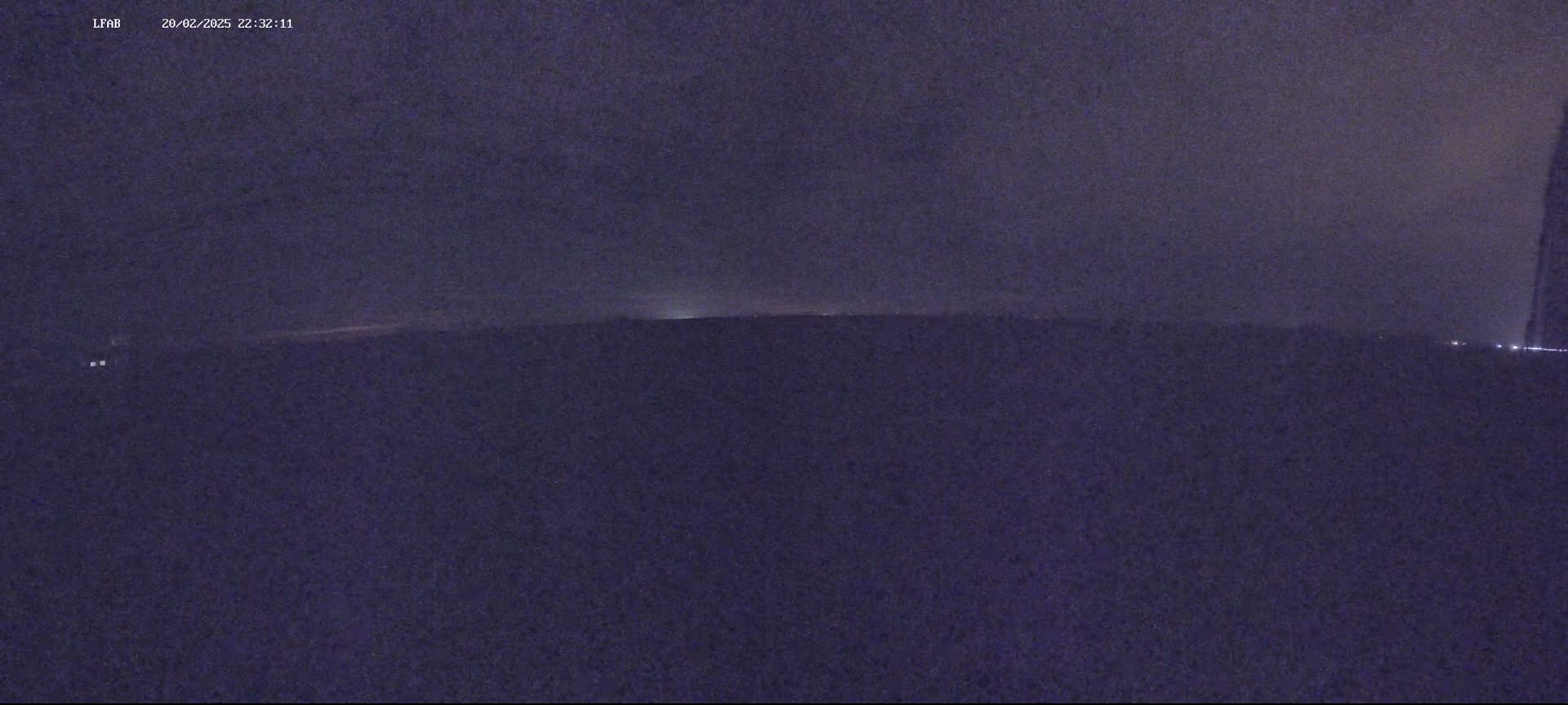 A dark, nighttime outdoor scene shows a flat, dark landscape under a mostly cloudy sky with a faint glow on the horizon.