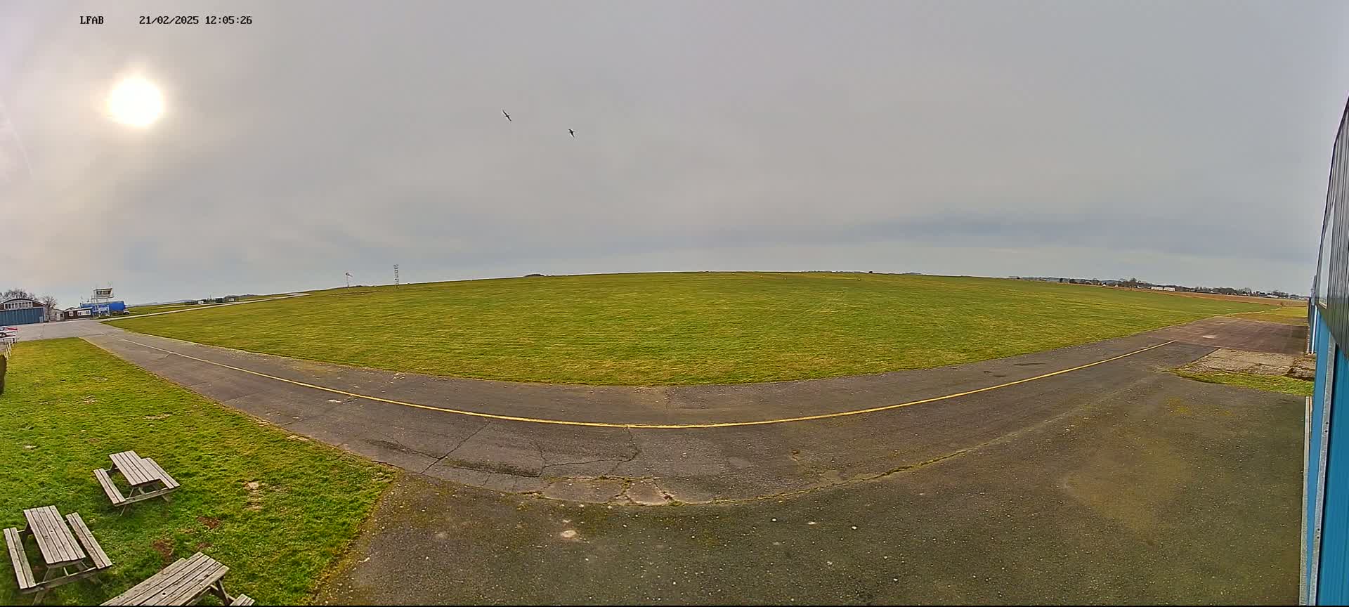 A paved path curves through a grassy airfield under a mostly cloudy sky with a bright sun.
