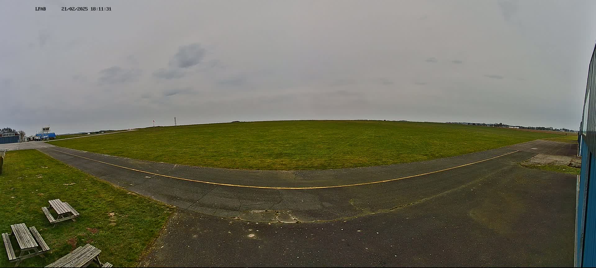 A grassy airfield with a paved taxiway and picnic tables is visible under an overcast sky.