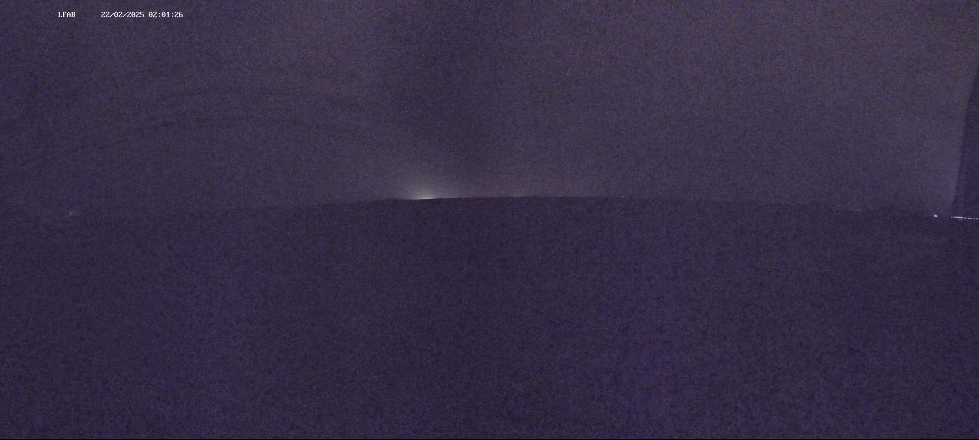 A dark, nighttime landscape is shown with a faint light source visible on the horizon under an overcast sky.