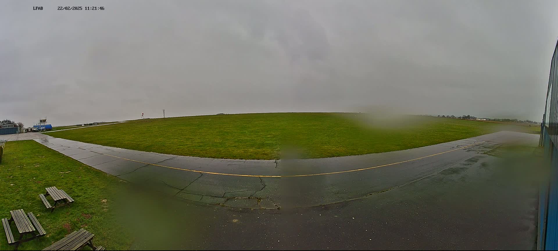 A wet, grassy airfield is seen on an overcast day, with a paved taxiway leading toward a hangar.