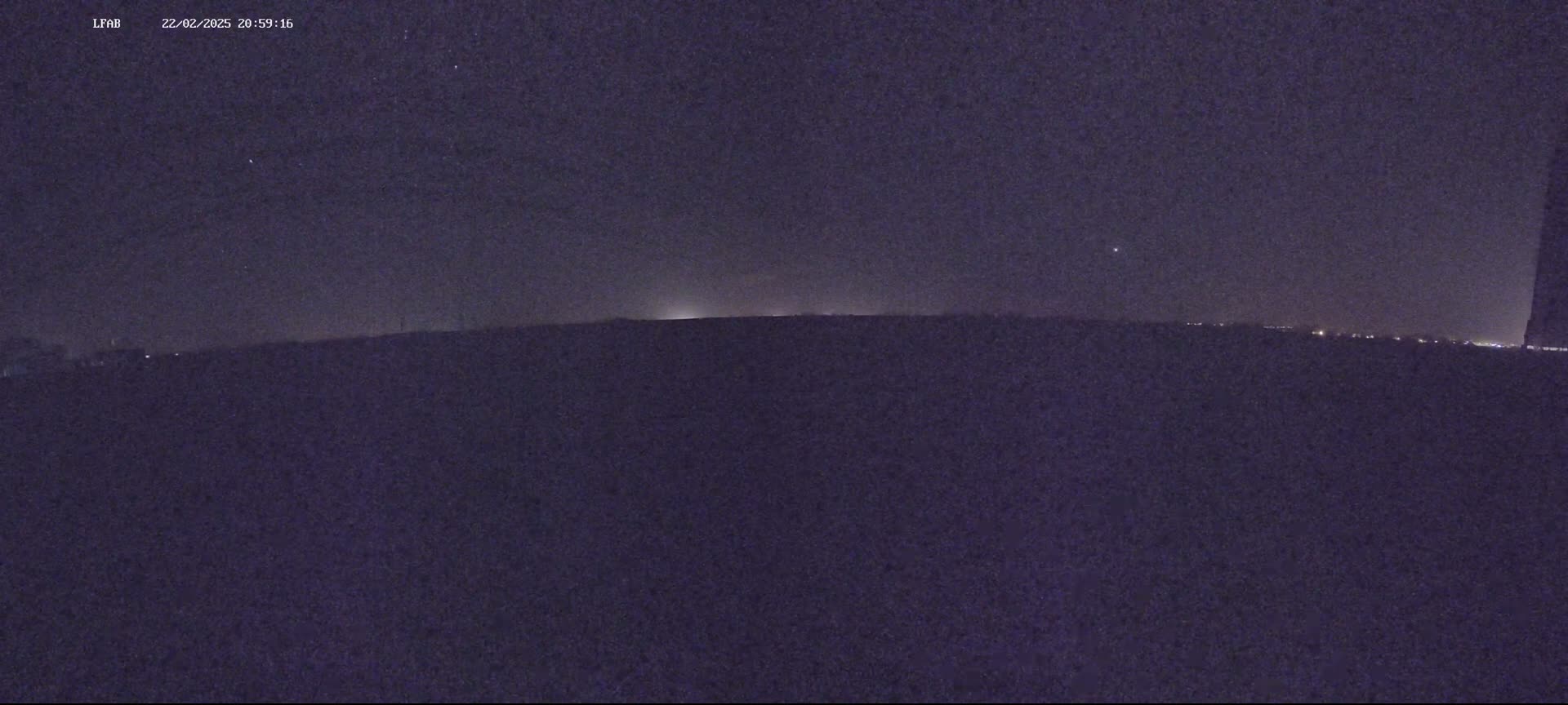 A dark, nighttime landscape shows a flat horizon line with a few distant lights under a mostly clear, starry sky.