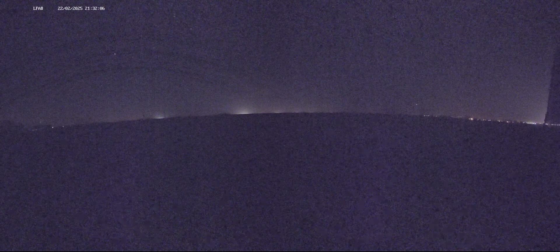 A dark, nighttime landscape shows a faint horizon line with distant lights under a mostly clear, starry sky.
