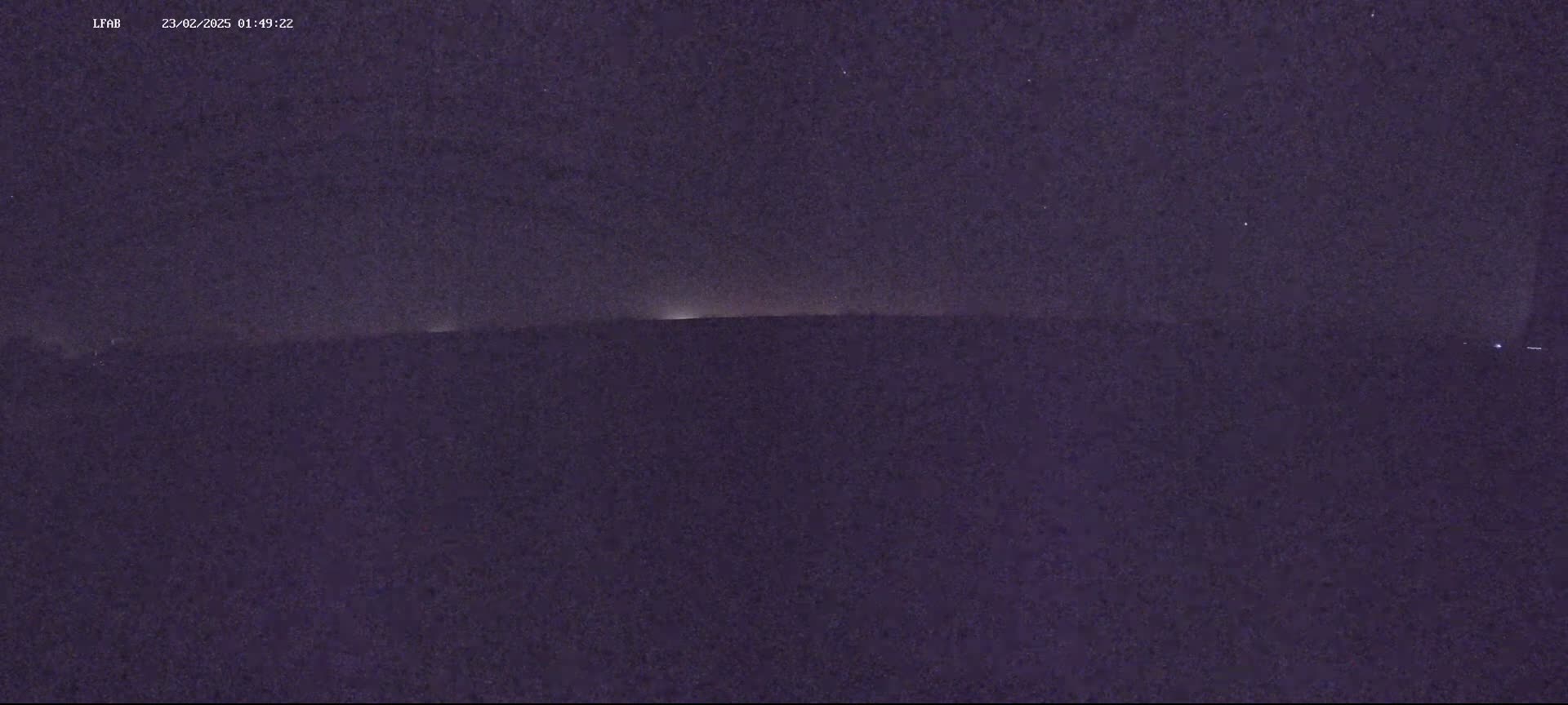 A dark, nighttime landscape shows a flat horizon line with a faint, low light source in the distance under a mostly clear, dark sky.