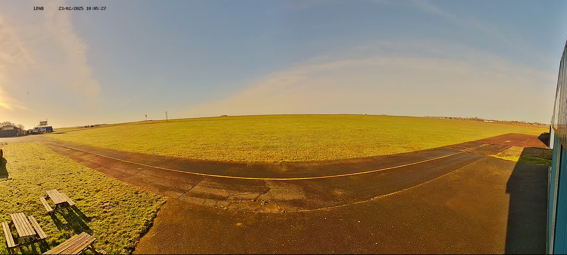 A grassy airfield under a mostly clear, sunny sky shows a paved taxiway leading to a large expanse of short, yellow-green grass.