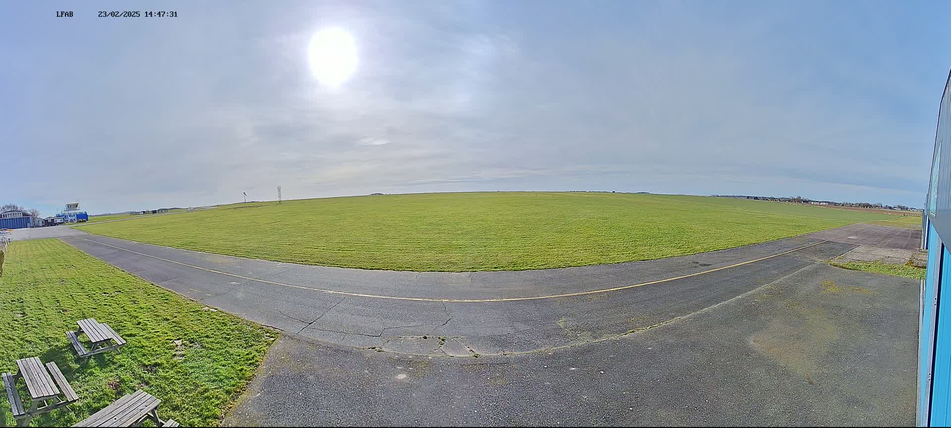 A sunny day reveals a grassy airfield with a paved taxiway curving from the foreground towards a distant hangar, under a mostly clear sky.