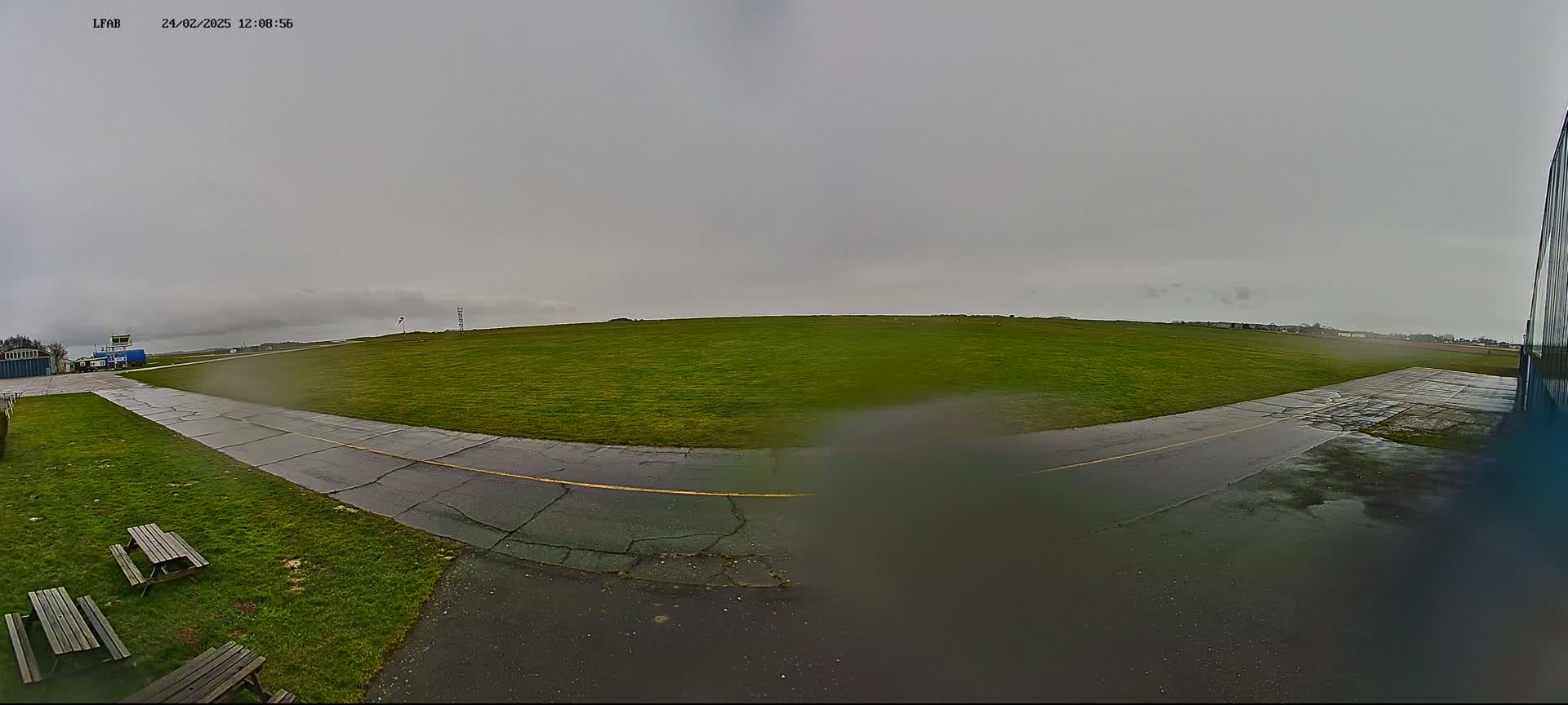 A wet airfield with a grassy field, a paved taxiway, and picnic tables is visible under an overcast sky.