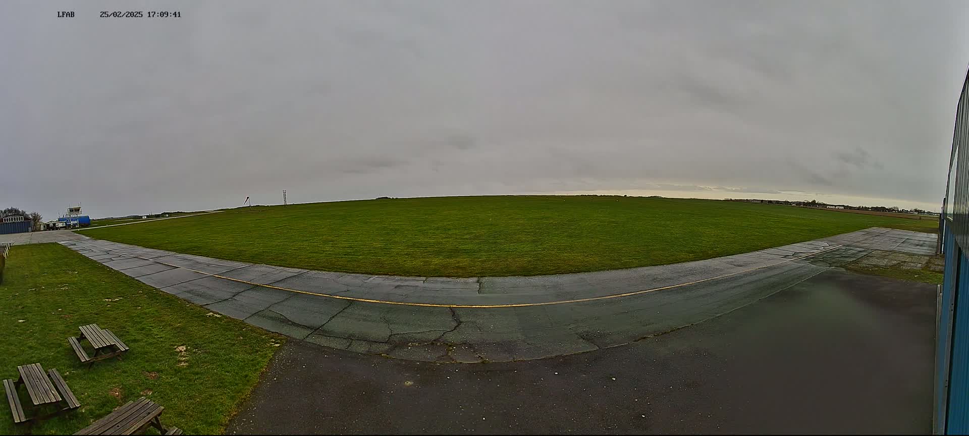 A grassy airfield with a paved taxiway curves toward a hangar under an overcast sky.