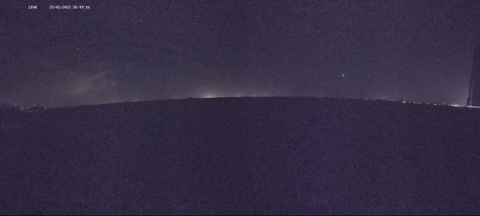 A dark, nighttime landscape shows a flat horizon line with faint lights in the distance under a mostly clear, starry sky.