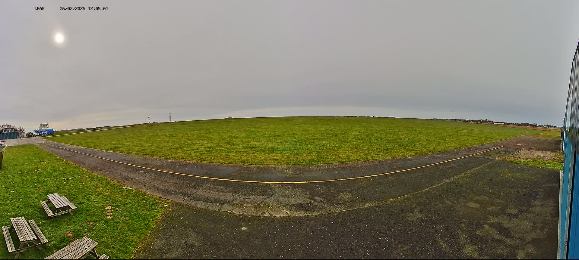 A wide, grassy airfield with a paved taxiway curves to the left under an overcast sky with a bright sun visible.