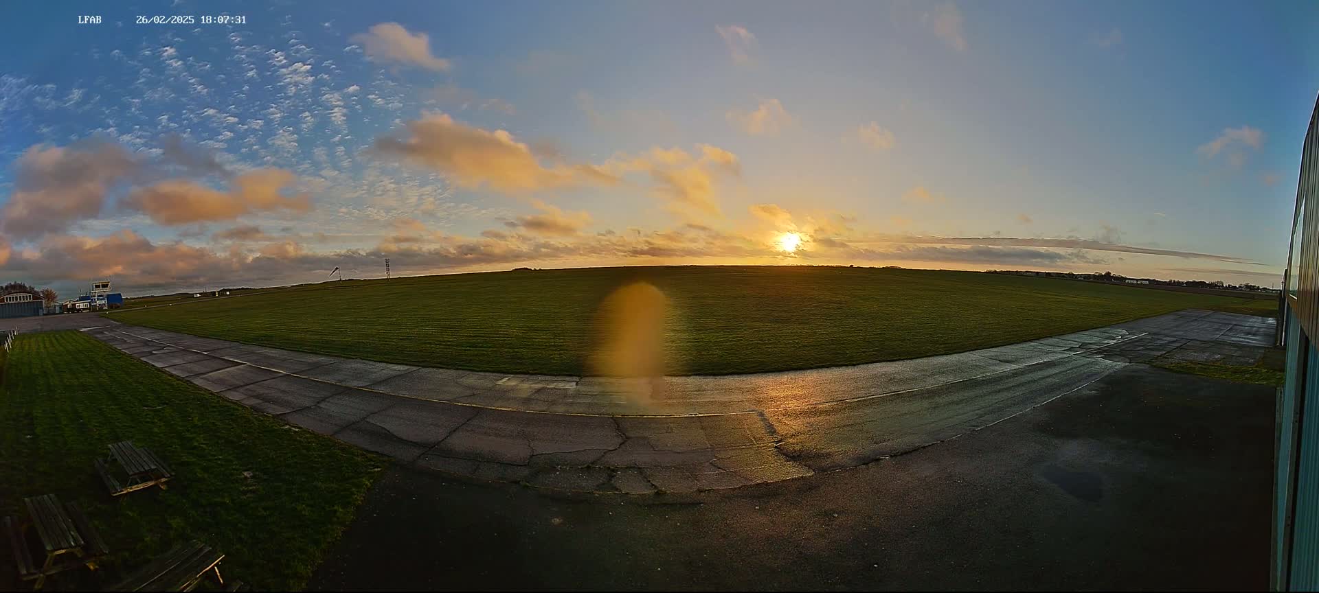 A grassy airfield with a paved taxiway is bathed in the golden light of a setting sun under a partly cloudy sky.