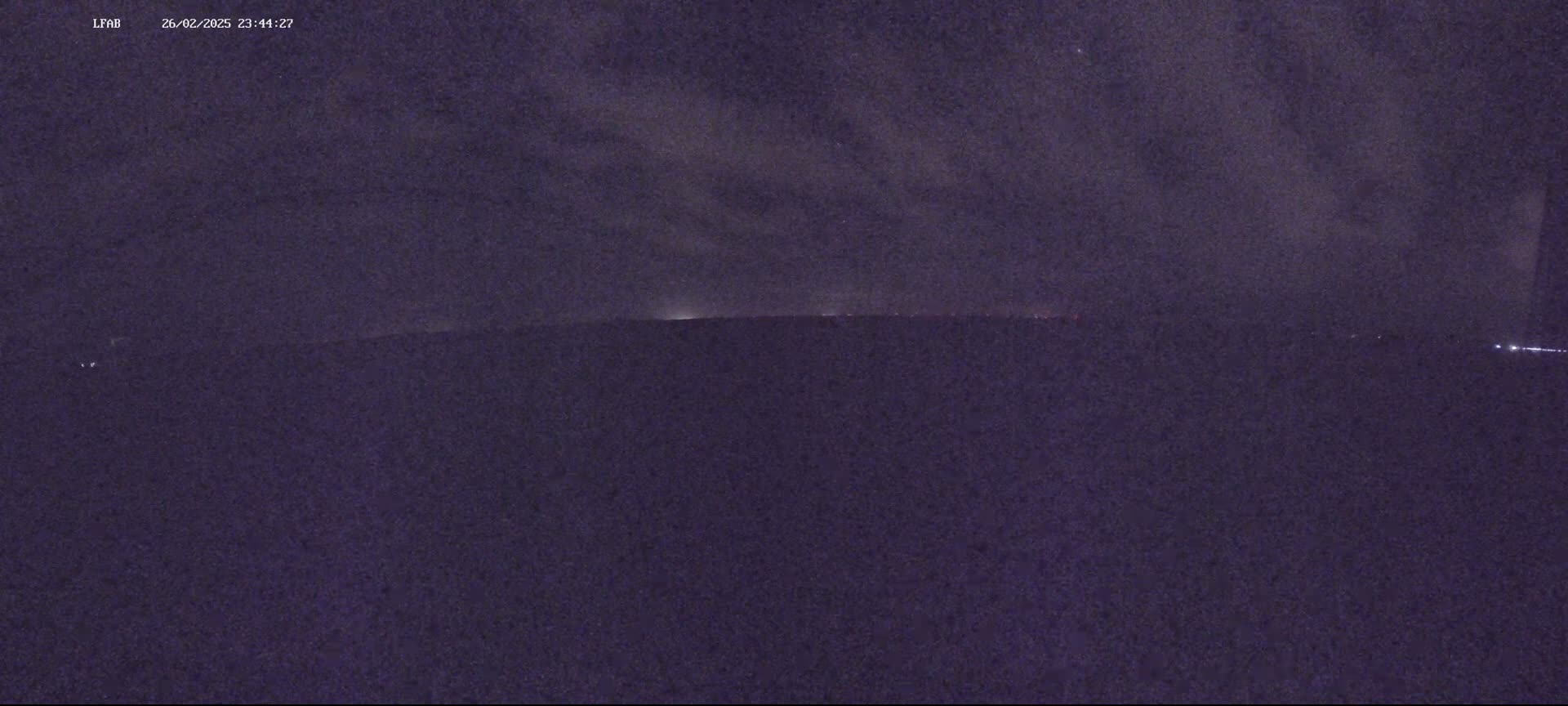 A nighttime view shows a dark landscape under a cloudy sky with faint lights on the horizon.