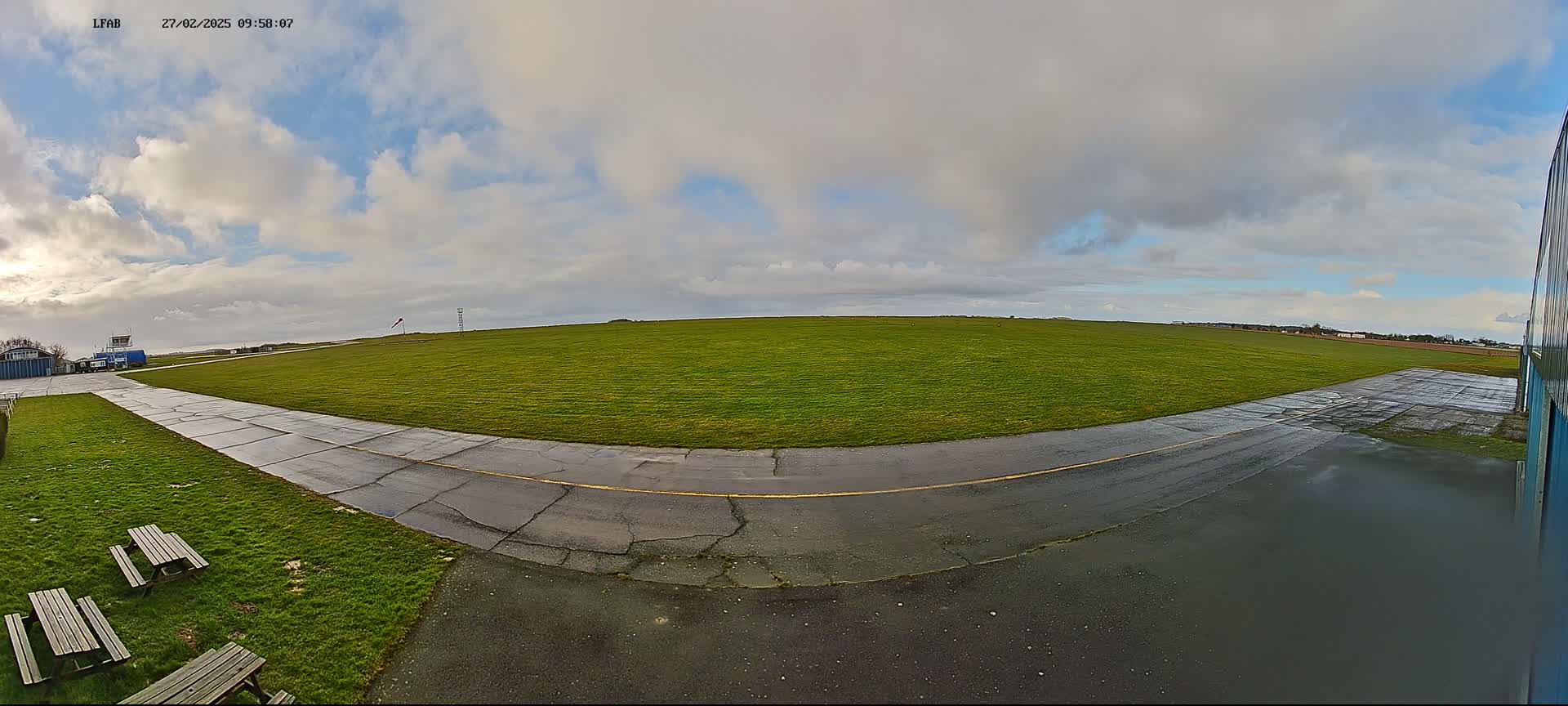 A grassy airfield with a paved taxiway curves toward a hangar under a partly cloudy sky.
