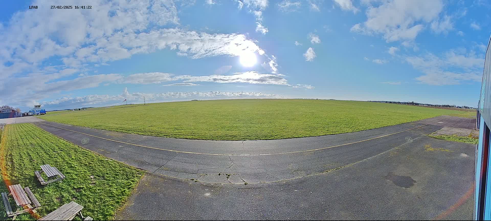 A wide, grassy airfield with a paved taxiway curves towards the viewer under a mostly sunny sky with scattered clouds.