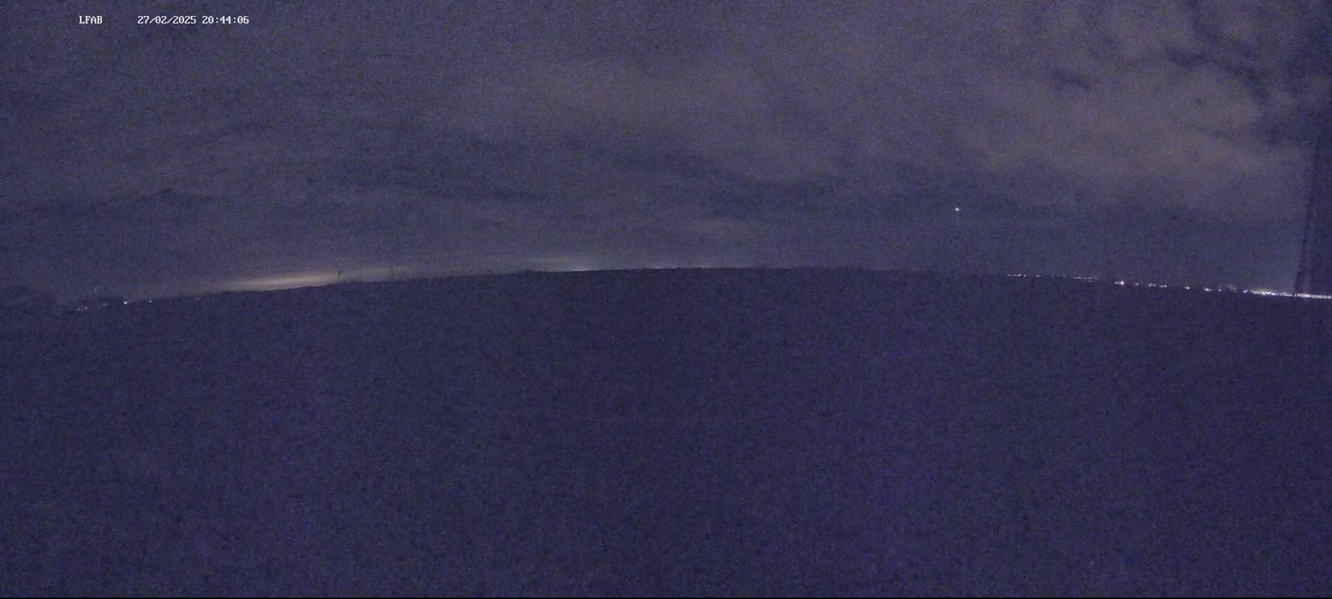 A nighttime, low-light view shows a dark landscape under a cloudy sky, with a faint horizon line showing distant lights.