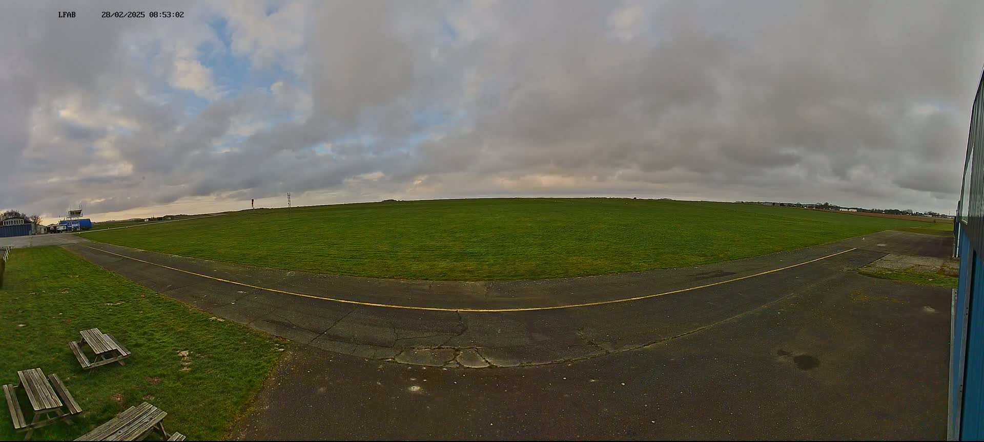 A grassy airfield under a cloudy sky features a paved taxiway curving towards a hangar, with picnic tables visible in the foreground.