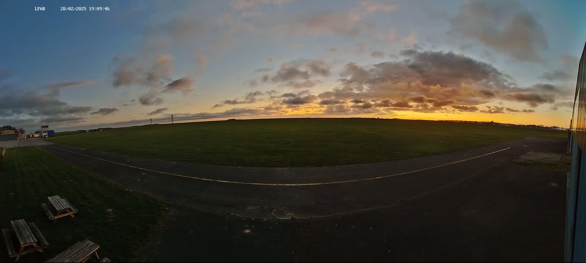 A grassy airfield with a paved taxiway is seen at sunset under a cloudy sky.