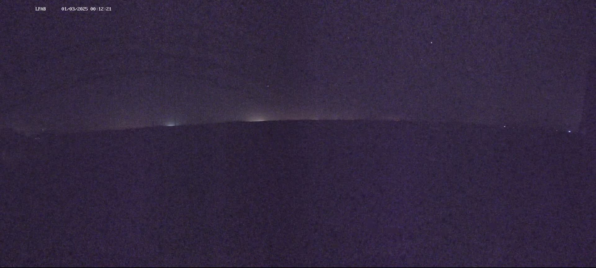 A dark, nighttime landscape shows a distant horizon line with faint lights visible against a mostly clear, starry sky.