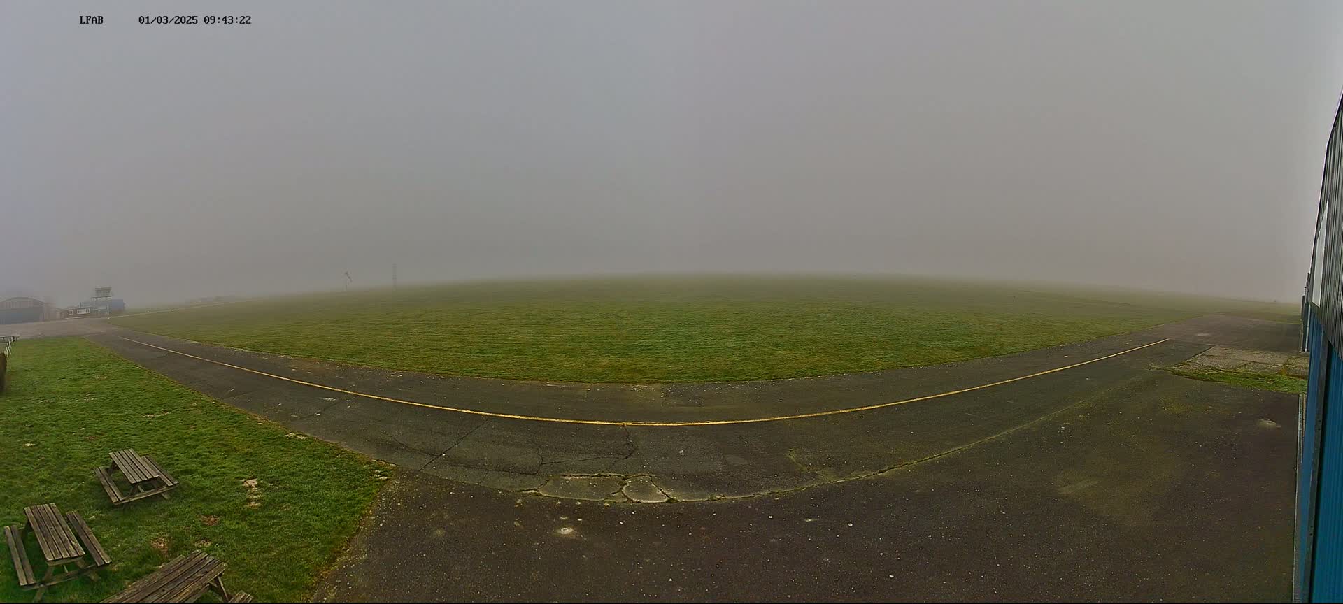 A grassy airfield is shrouded in thick fog, with a paved taxiway and a few picnic tables visible in the foreground.