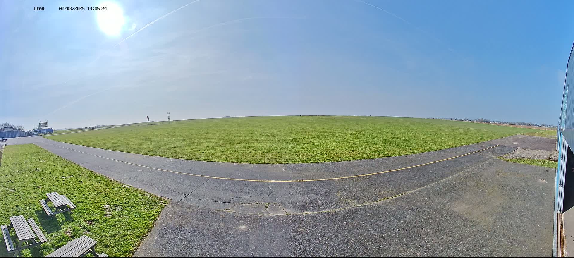 A grassy airfield under a sunny, clear sky is seen from a ground-level perspective, with a paved taxiway curving toward the viewer.