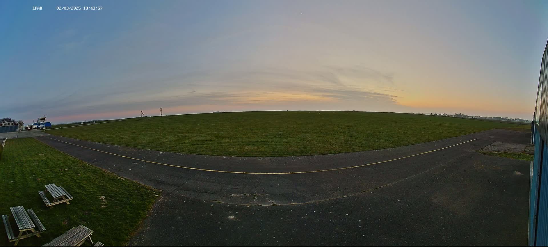 A grassy airfield at sunset, with a paved taxiway curving towards a small building, and picnic tables nearby.