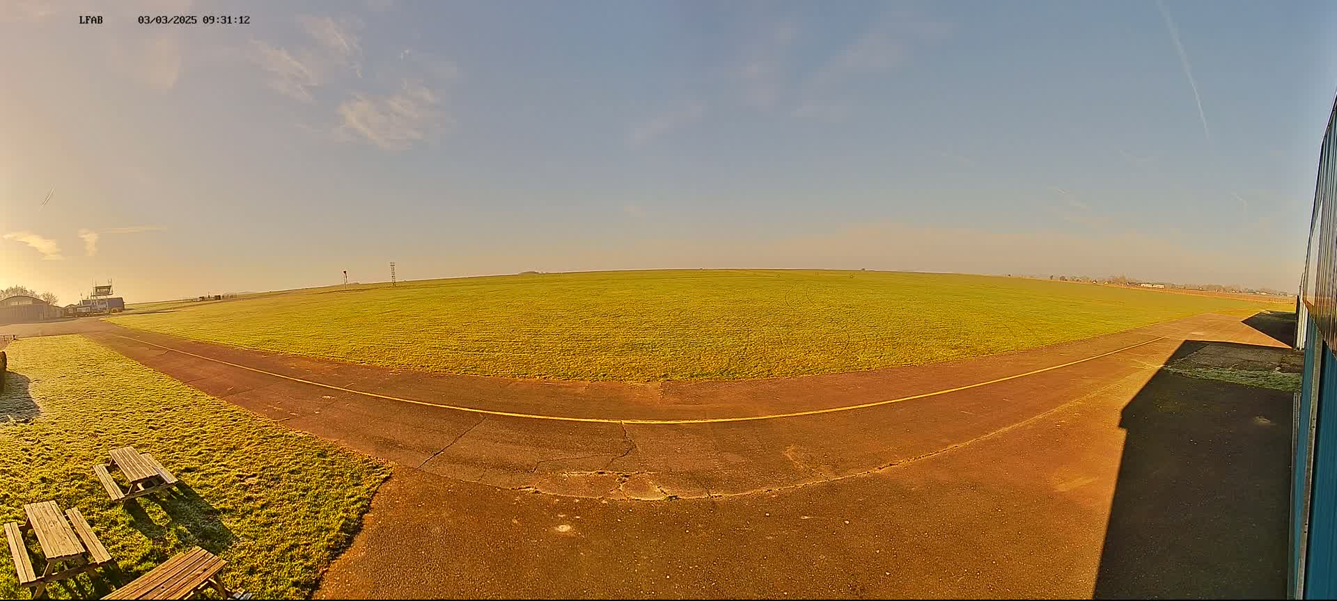 A grassy airfield with a paved runway and picnic tables is visible under a mostly clear, sunny sky.