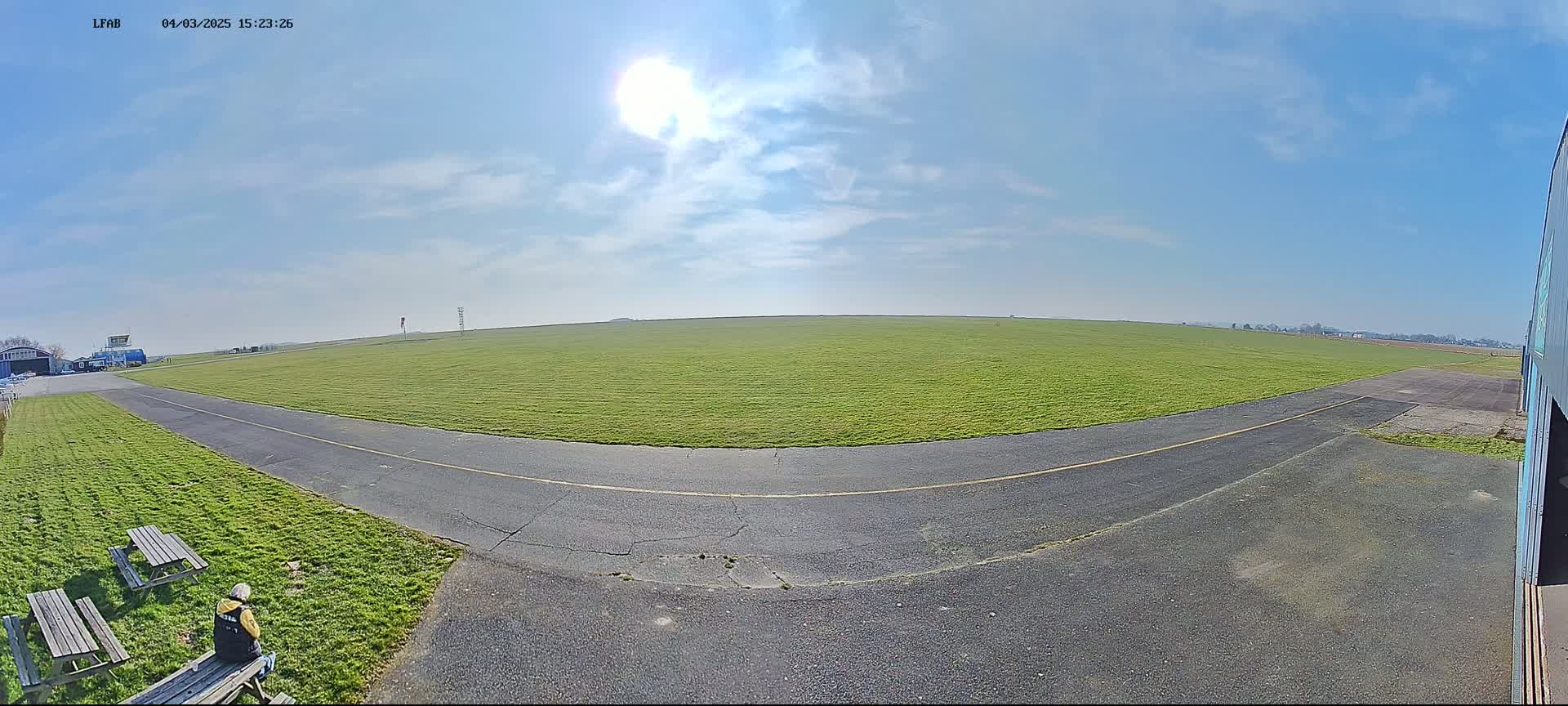A sunny day reveals a grassy airfield with a paved taxiway curving towards a hangar, picnic tables, and a person sitting at one.