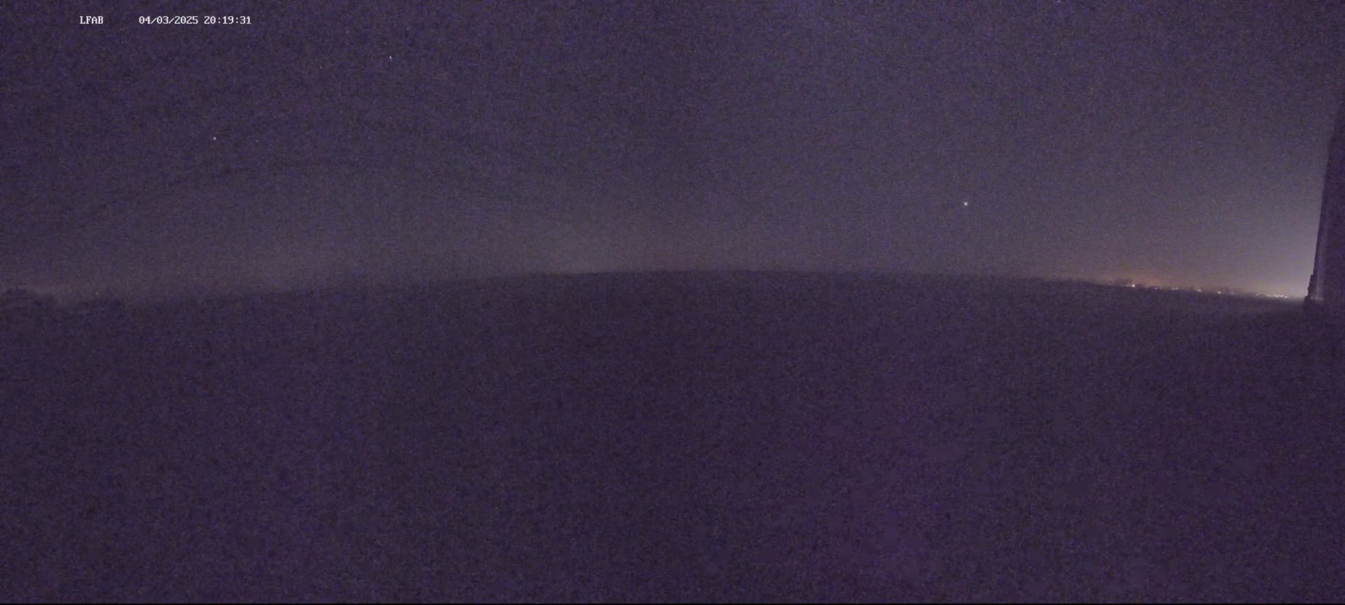 A dark, night-time view shows a flat, dark landscape under a mostly clear, starry sky.