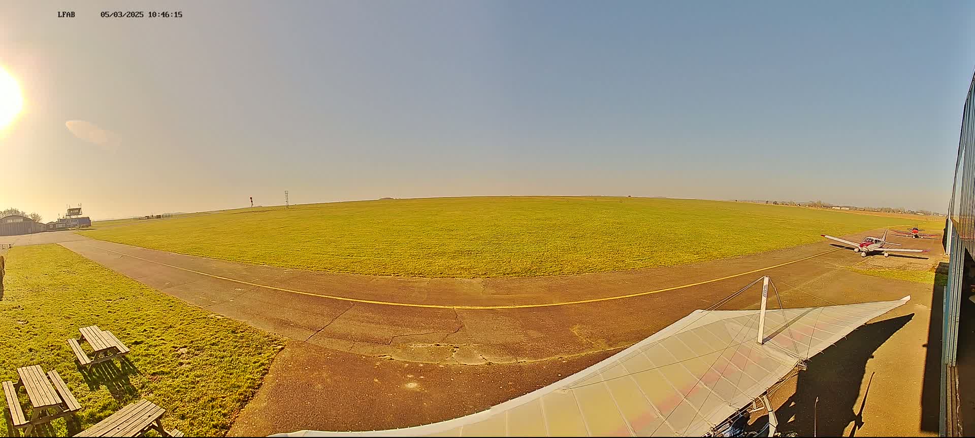 A sunny day at a small airfield shows a grassy runway with several small airplanes parked and a hang glider wing visible in the foreground.