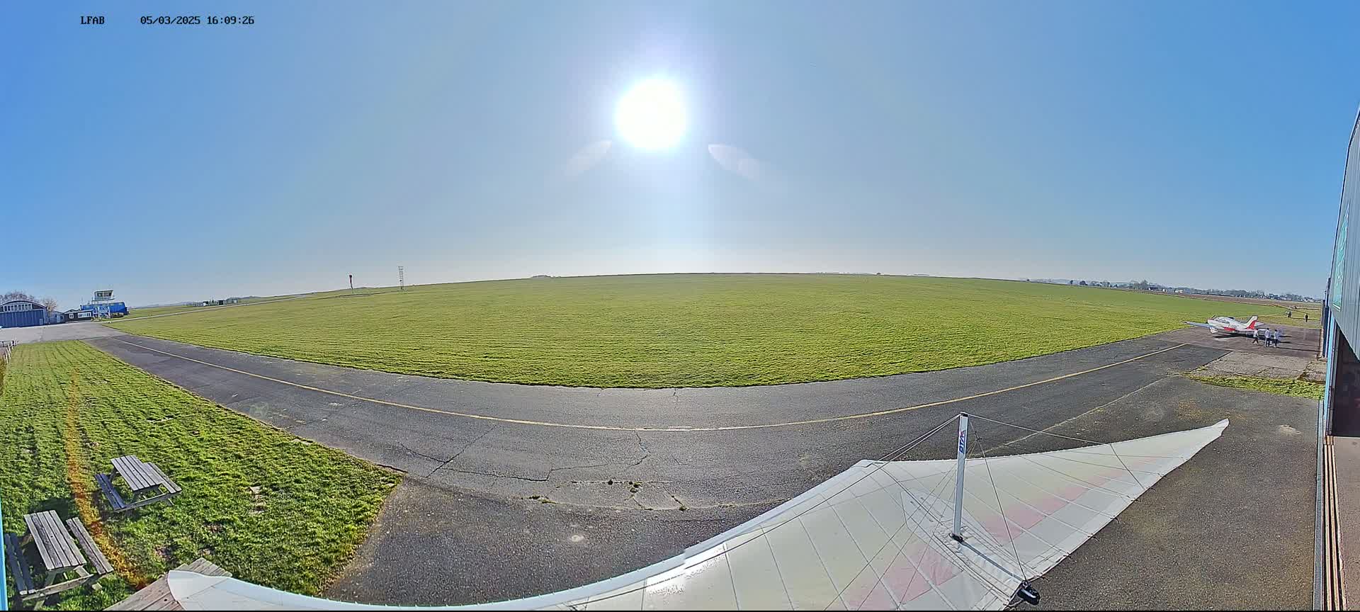 A sunny day at a grass airfield shows a small airplane parked near a hangar, with a hang glider's wing in the foreground and a paved taxiway curving through a grassy field.