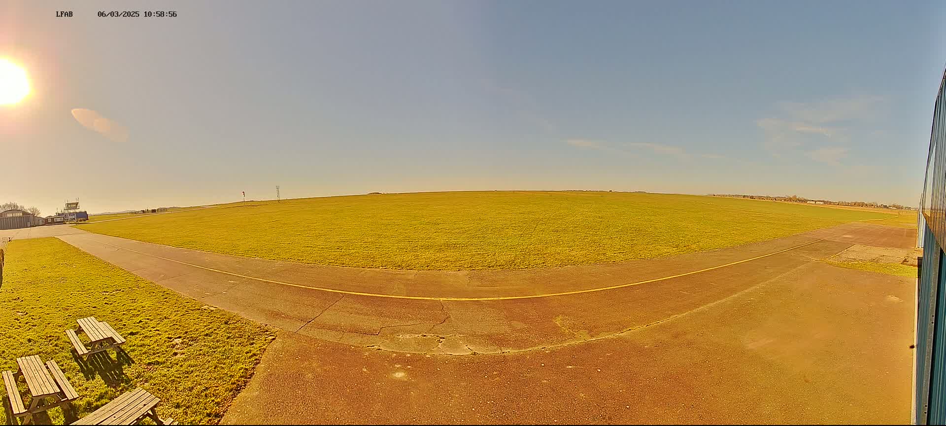 A sunny day at a grassy airfield shows a paved taxiway leading to a large, flat field with picnic tables visible in the foreground.