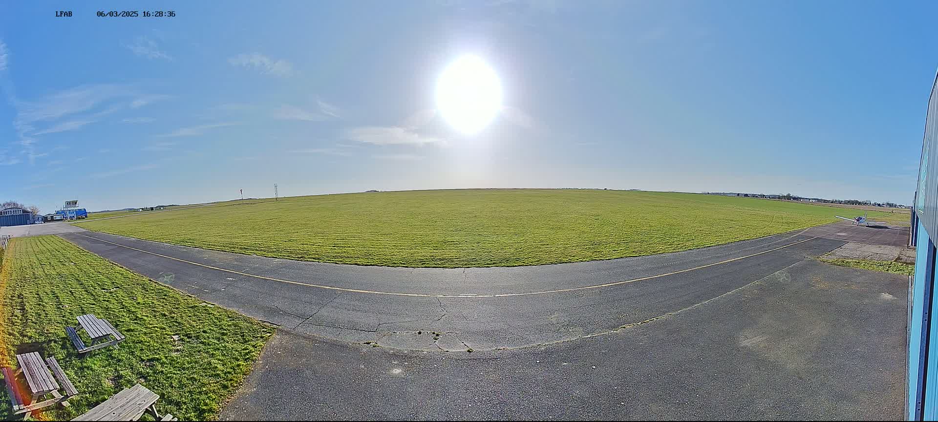 A sunny day at a small airfield shows a grassy field, a paved runway, and a small airplane on the tarmac.