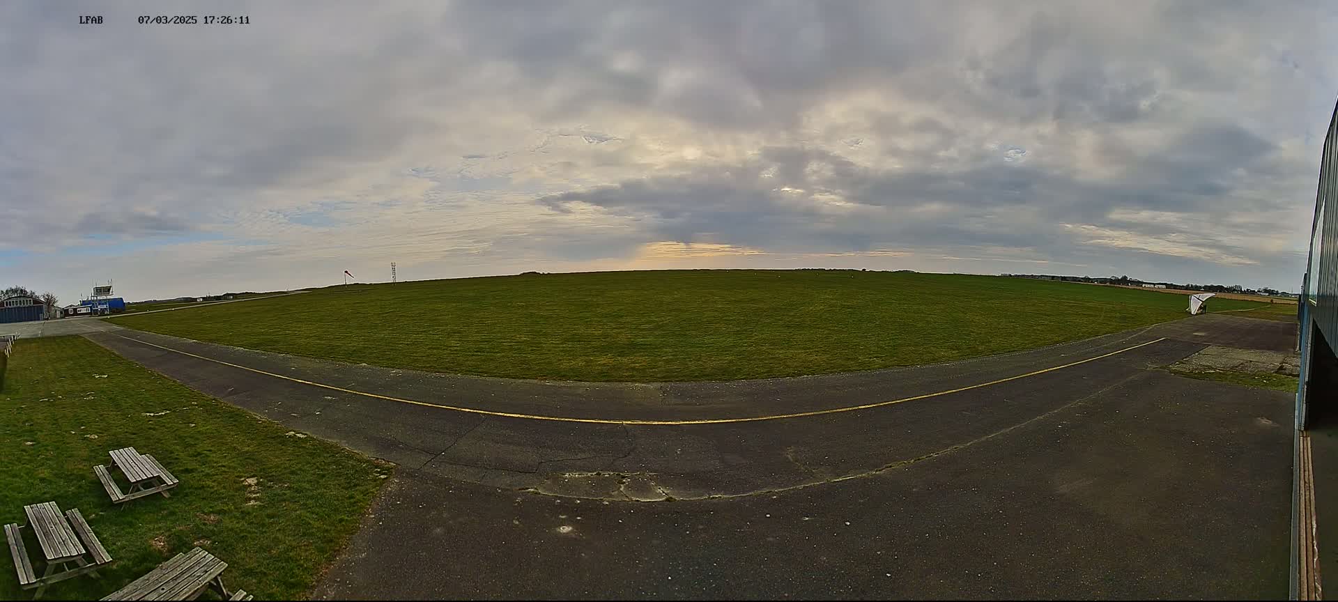 A grassy airfield with a paved taxiway and picnic tables under a cloudy sky.