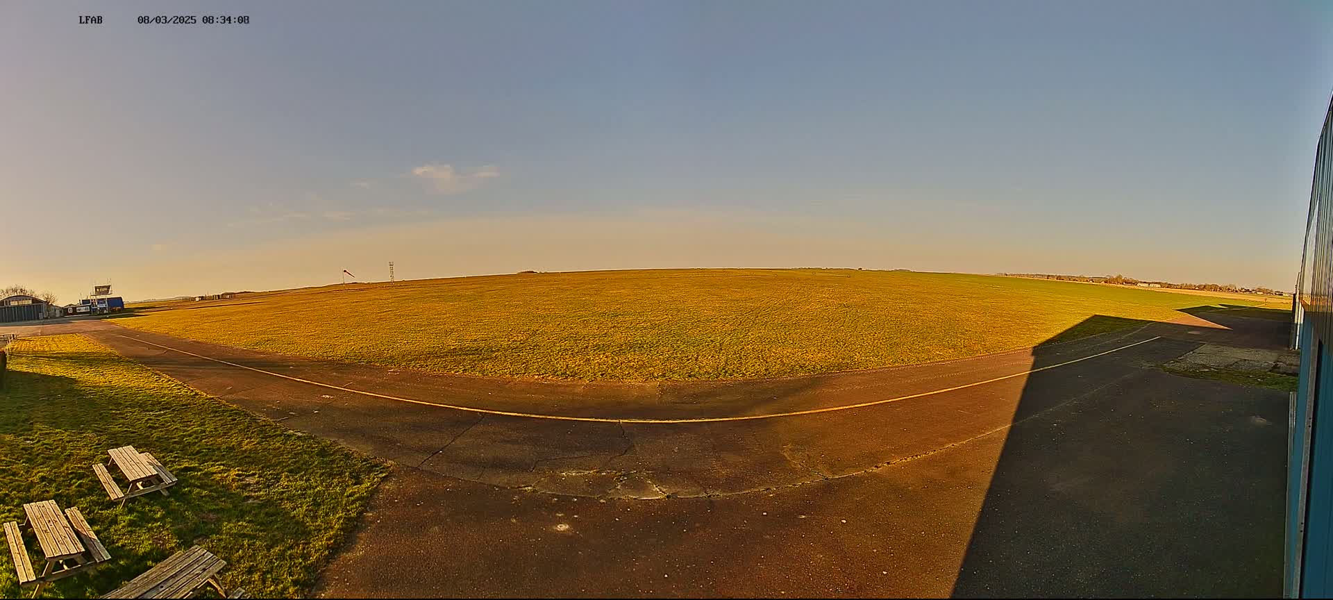 A wide, grassy airfield with a paved taxiway curves toward a hangar under a clear, bright sky.
