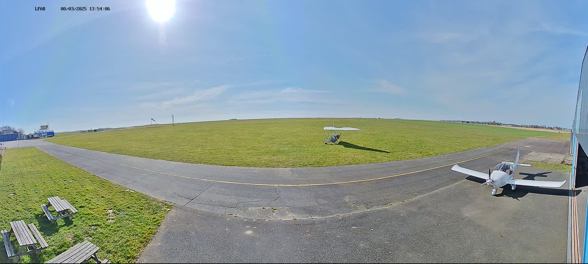 A sunny day at a grass airfield shows a small airplane parked near a hangar, with a hang glider resting on the grass further down the runway.