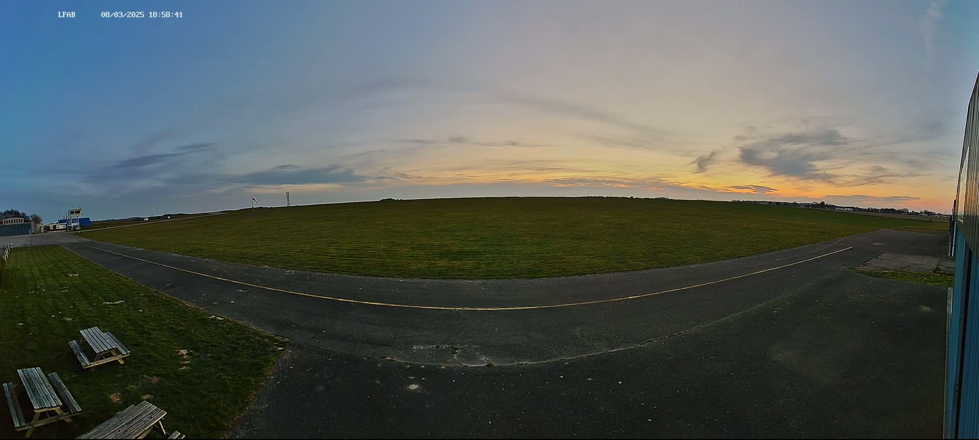 A grassy airfield at sunset, with a paved taxiway curving toward the viewer, and picnic tables nearby.