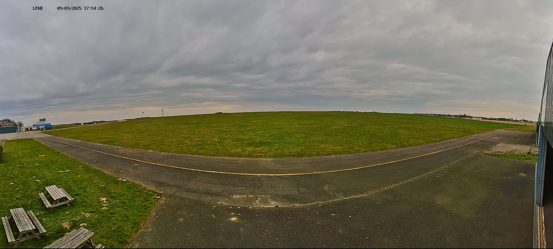 A grassy airfield with a paved taxiway, picnic tables, and a building under an overcast sky.
