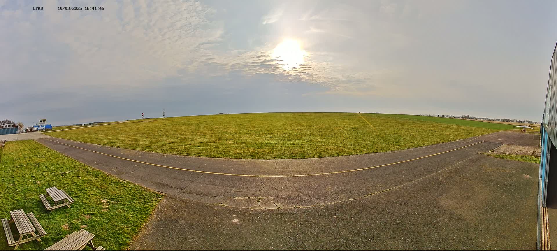 A grassy airfield with a paved runway and picnic tables is visible under a partly cloudy sky with the sun shining brightly.
